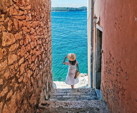 A person in a white dress and sun hat descends stone steps between rustic walls toward a bright blue sea, evoking a sense of serene adventure.