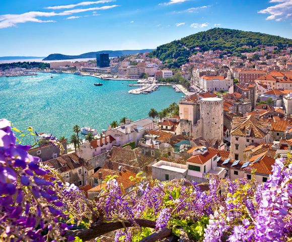 Scenic view of a coastal town with terracotta rooftops and historic stone buildings. The turquoise sea sparkles under the sun, surrounded by green hills and purple flowers in the foreground.