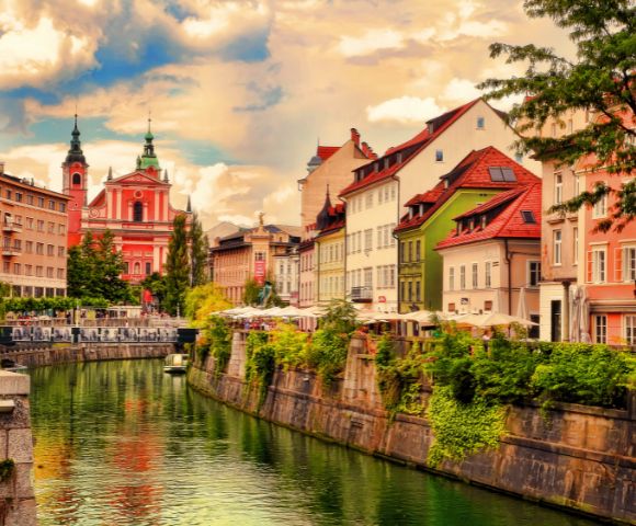 Colorful riverside buildings line a peaceful canal in Ljubljana. The sky is partly cloudy, and a prominent pink church stands in the background.