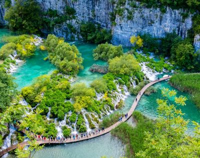 Vibrant aerial view of Plitvice Lakes, Croatia, featuring turquoise waters, lush green foliage, and a winding wooden boardwalk with visitors exploring.