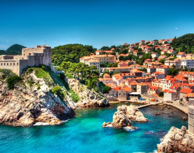 Coastal cityscape with a historic fortress atop a rocky cliff, overlooking turquoise waters. Nearby, houses with red-tiled roofs under a clear blue sky.