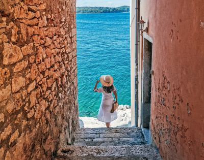 A person in a white dress and sun hat descends stone steps between rustic walls toward a bright blue sea, evoking a sense of serene adventure.