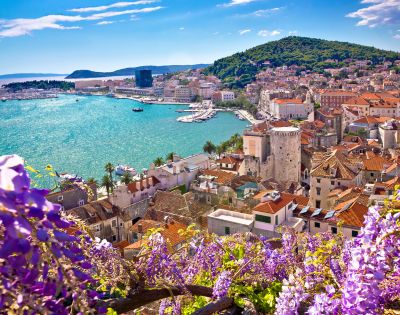 Scenic view of a coastal town with terracotta rooftops and historic stone buildings. The turquoise sea sparkles under the sun, surrounded by green hills and purple flowers in the foreground.