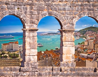View of a coastal town from a stone wall with three arches. Turquoise sea, boats, and red-tiled roofs under a vibrant blue sky create a serene scene.
