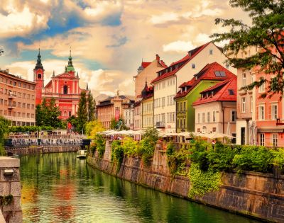Colorful riverside buildings line a peaceful canal in Ljubljana. The sky is partly cloudy, and a prominent pink church stands in the background.