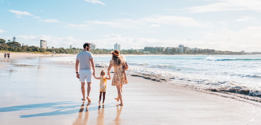 A family walks hand-in-hand along a sunny beach, leaving footprints in the wet sand. The sky is clear, and buildings are visible in the distance.