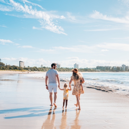 A family walks hand-in-hand along a sunny beach, leaving footprints in the wet sand. The sky is clear, and buildings are visible in the distance.