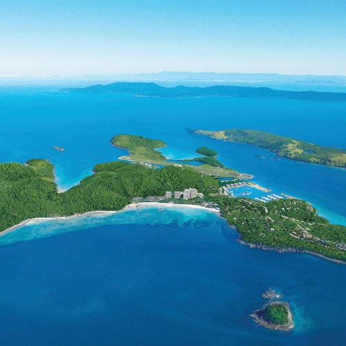 Aerial view of a lush, green island surrounded by vibrant blue sea. Small buildings are nestled among trees, with clear skies imparting a serene, tropical vibe.
