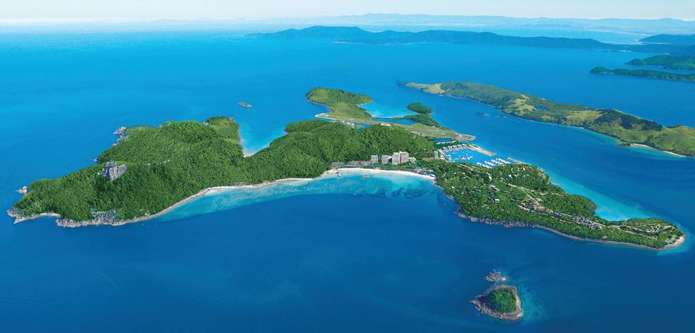 Aerial view of a lush, green island surrounded by vibrant blue sea. Small buildings are nestled among trees, with clear skies imparting a serene, tropical vibe.