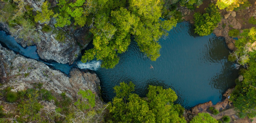 Aerial view of a tranquil blue lagoon surrounded by lush green trees and rocky terrain, with a person swimming in the center.
