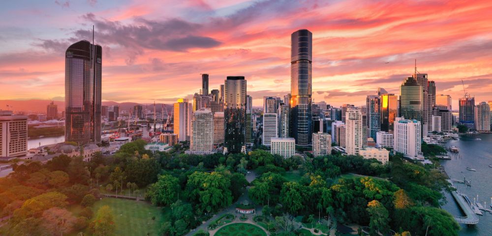 A vibrant city skyline at sunset with tall skyscrapers, lush green parks in the foreground, and a river on the right, under pink and orange clouds.