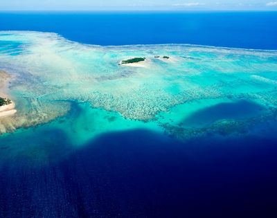 Aerial view of a vibrant turquoise ocean surrounding small, lush islands and coral reefs under a bright blue sky.