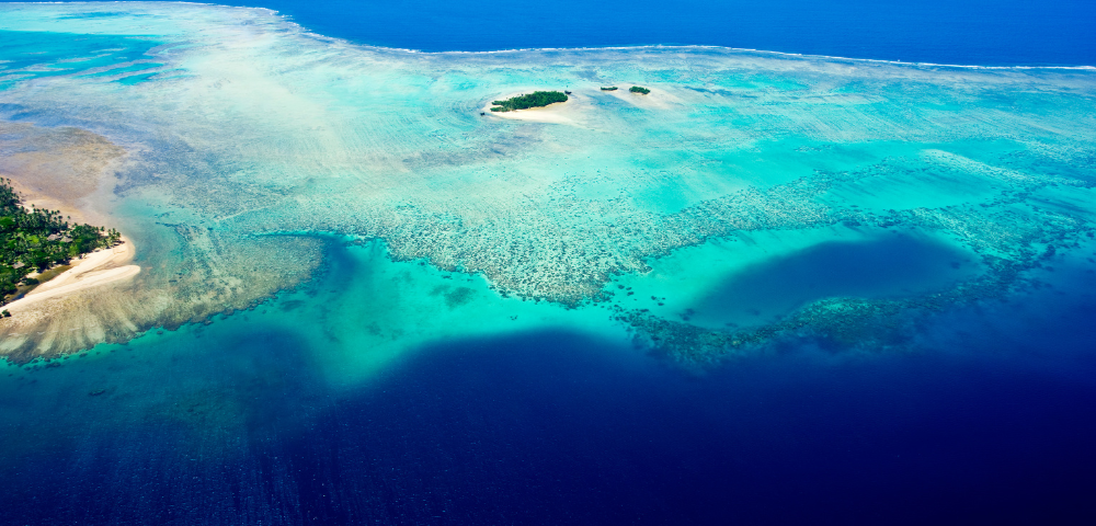 Aerial view of a vibrant turquoise ocean surrounding small, lush islands and coral reefs under a bright blue sky.