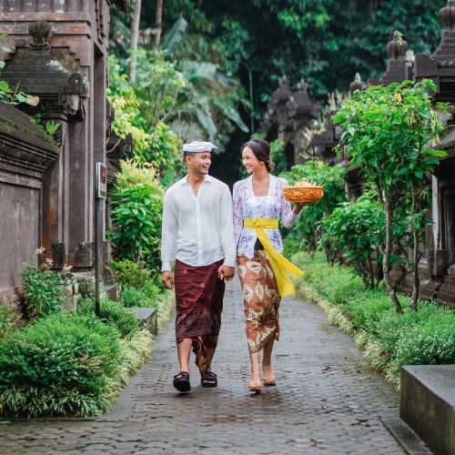 A couple in traditional Balinese attire walks hand-in-hand along a lush, stone pathway. They smile warmly, conveying a sense of joy and serenity.