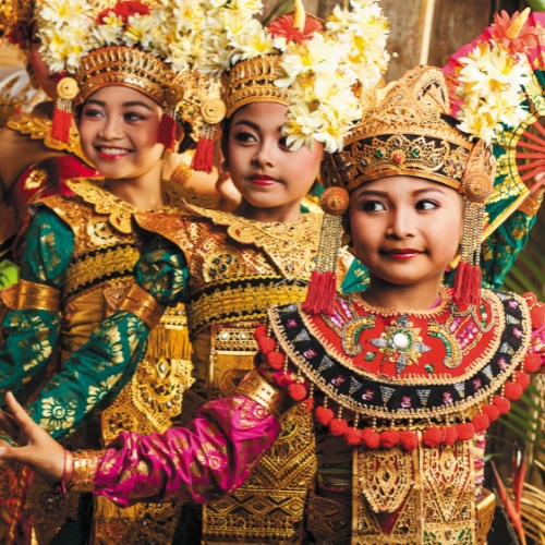 Three girls in vibrant Balinese costumes dance gracefully. They wear ornate headdresses adorned with flowers, conveying tradition.