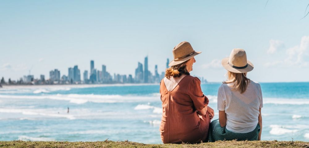 Two women in sun hats sit on grass by the ocean, overlooking waves and a city skyline under a clear blue sky.