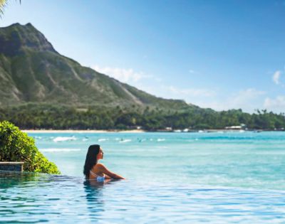 A serene infinity pool overlooking a turquoise ocean, with a backdrop of lush mountains and a clear blue sky.