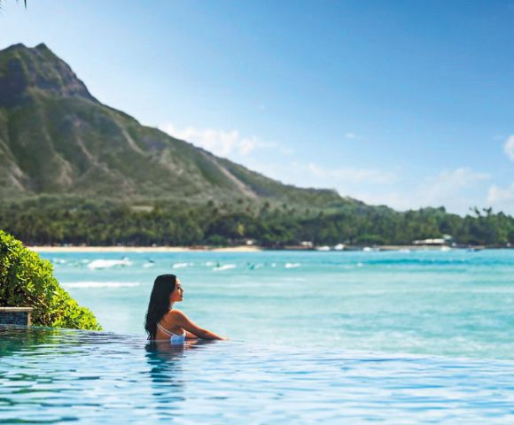 A serene infinity pool overlooking a turquoise ocean, with a backdrop of lush mountains and a clear blue sky.