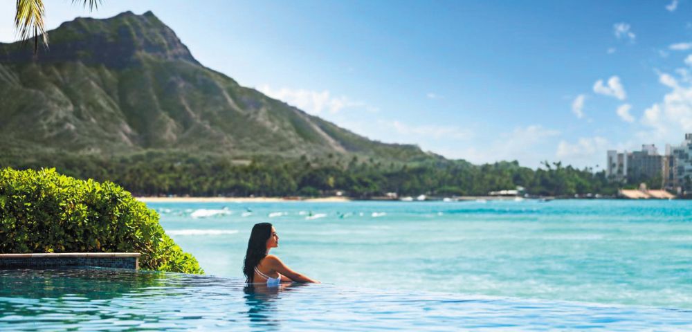 A serene infinity pool overlooking a turquoise ocean, with a backdrop of lush mountains and a clear blue sky.
