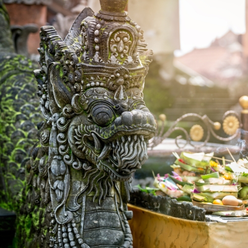 Ornate stone dragon statue with intricate carvings stands beside a colorful offering of flowers and fruit in woven leaf bowls.