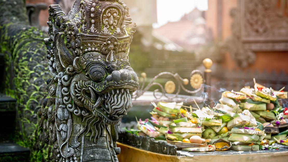 Ornate stone dragon statue with intricate carvings stands beside a colorful offering of flowers and fruit in woven leaf bowls.