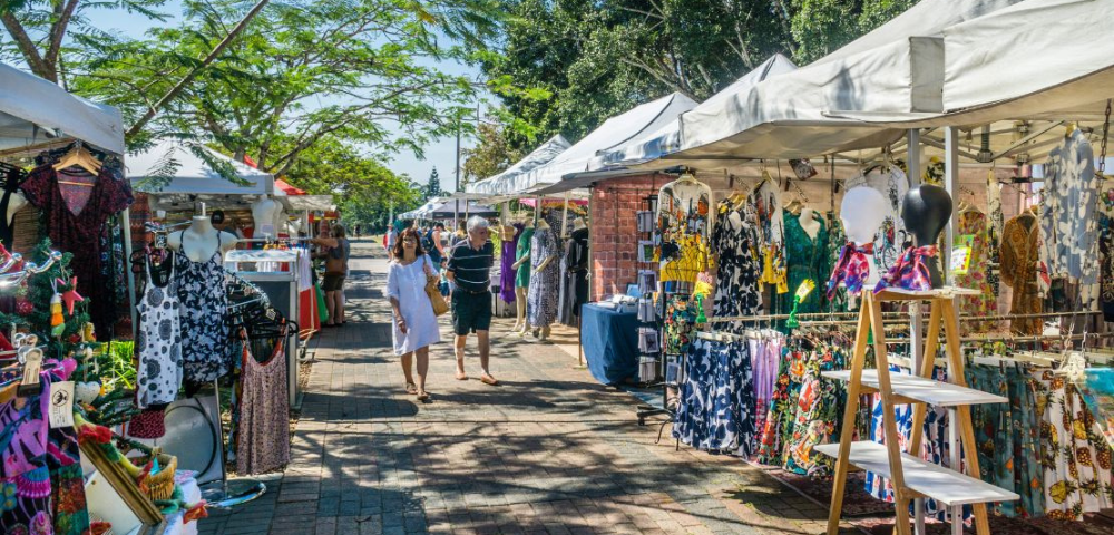 A vibrant outdoor market scene with stalls showcasing colorful clothing and accessories, as shoppers stroll along a sunny path.