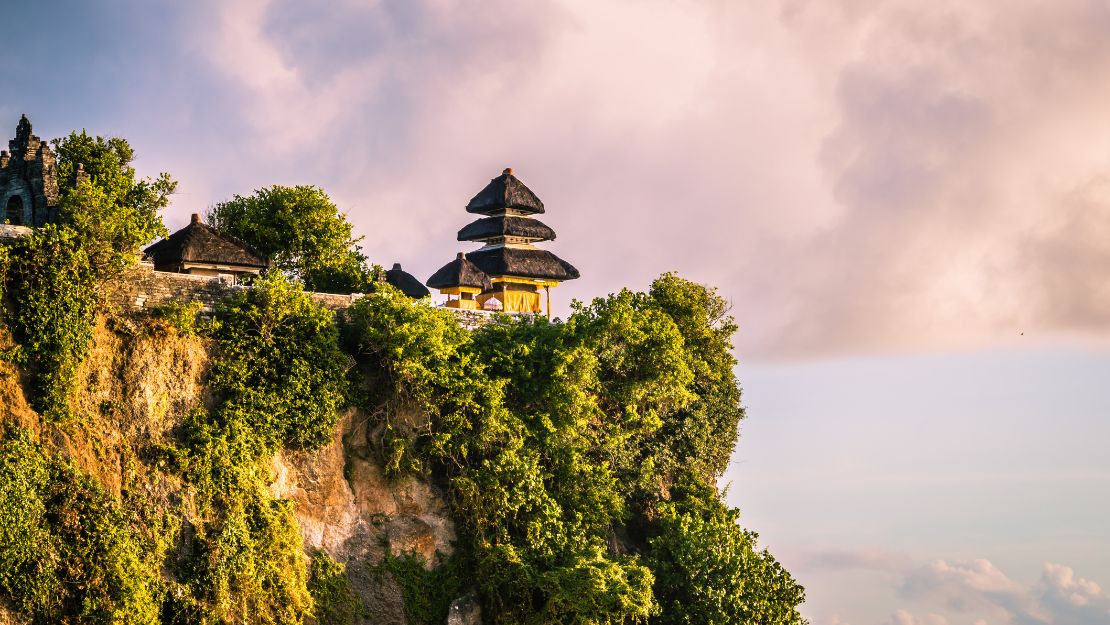 Clifftop temple surrounded by lush greenery under a pink-orange sky during sunset.