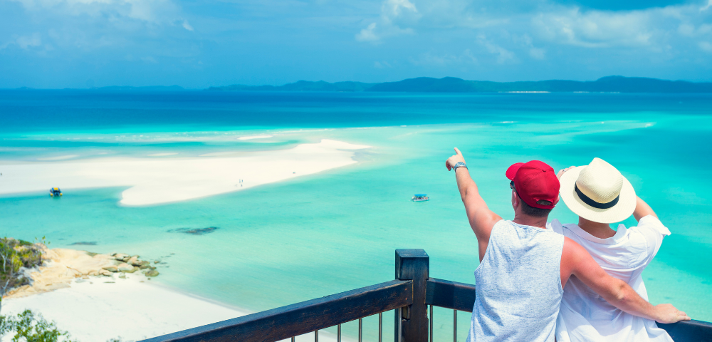 A couple stands together, gazing at a stunning azure seascape, pointing towards a sandy beach and small boats in the distance.