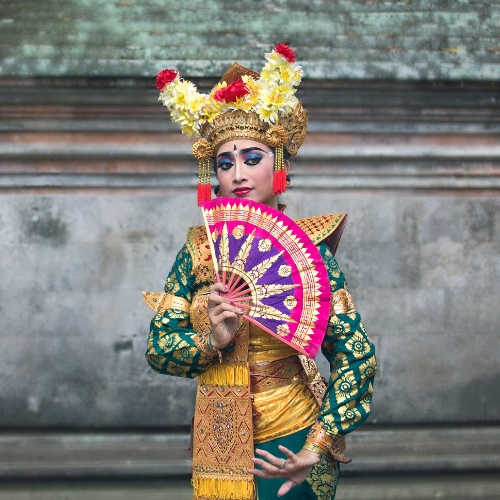 A person in traditional Balinese attire poses against a stone wall. They hold a vibrant pink fan, wearing an ornate headdress adorned with flowers.