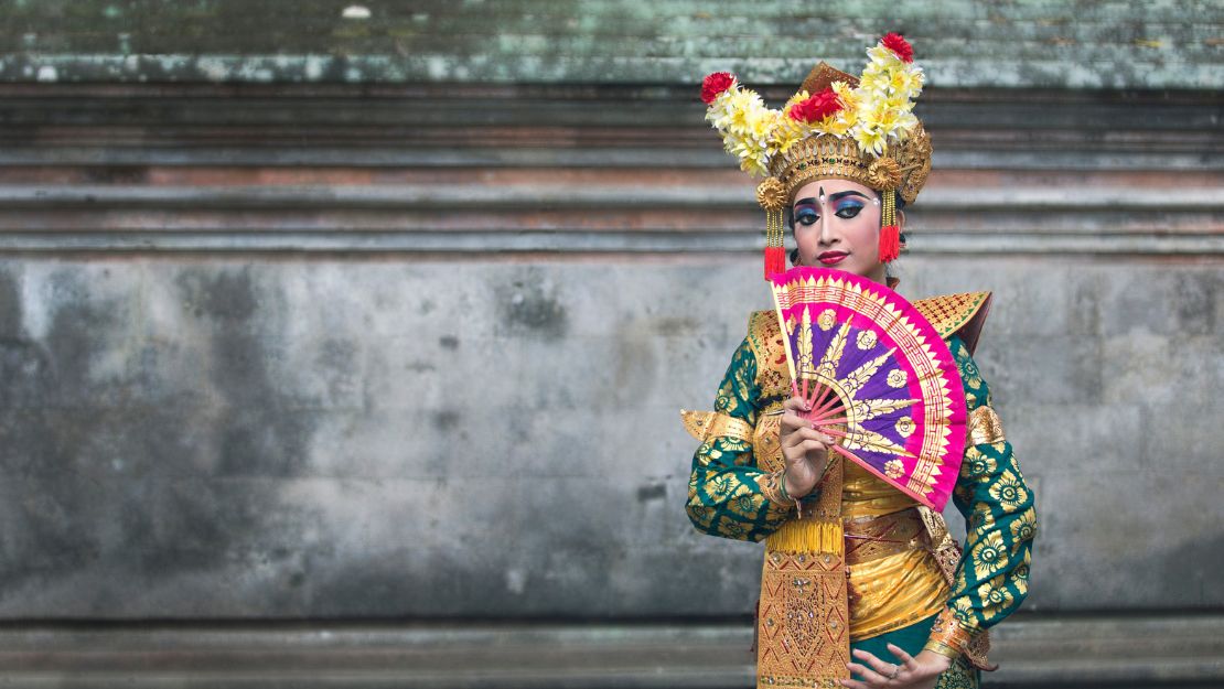 A person in traditional Balinese attire poses against a stone wall. They hold a vibrant pink fan, wearing an ornate headdress adorned with flowers.