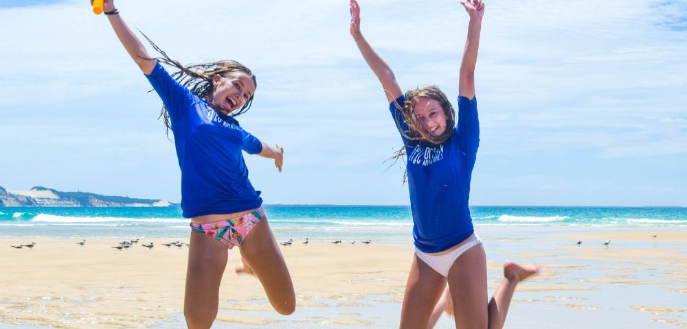Two girls in blue shirts leap joyfully on a sunny beach, with a vibrant ocean and sandy shore in the background.