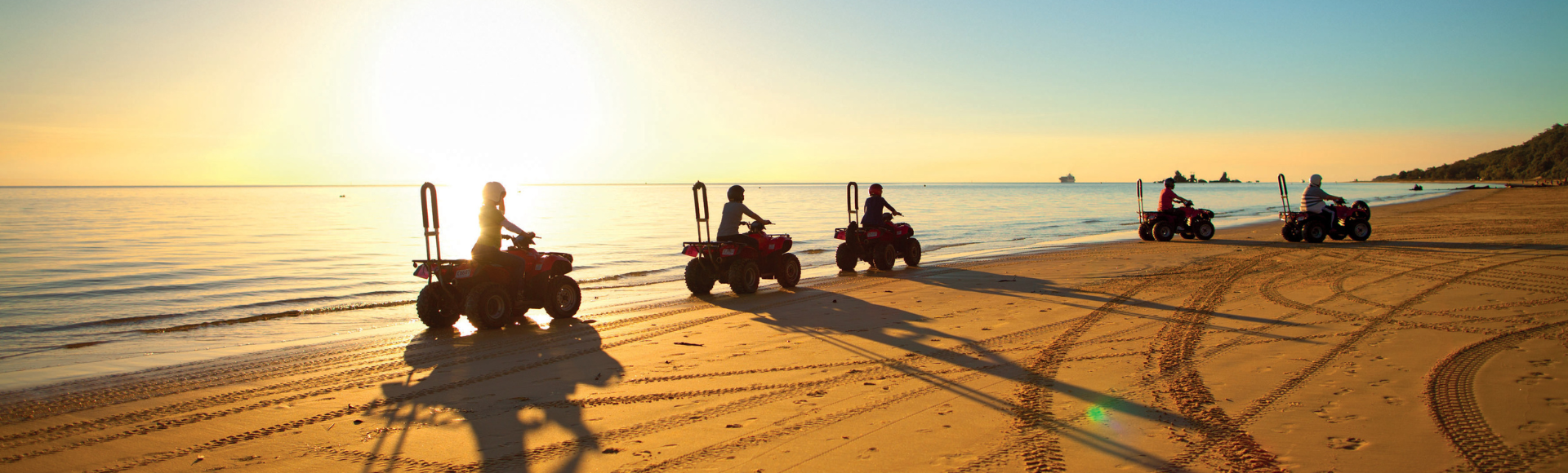 Silhouetted riders on ATVs cruising along a sandy beach at sunset, with calm water and a distant ship visible on the horizon.
