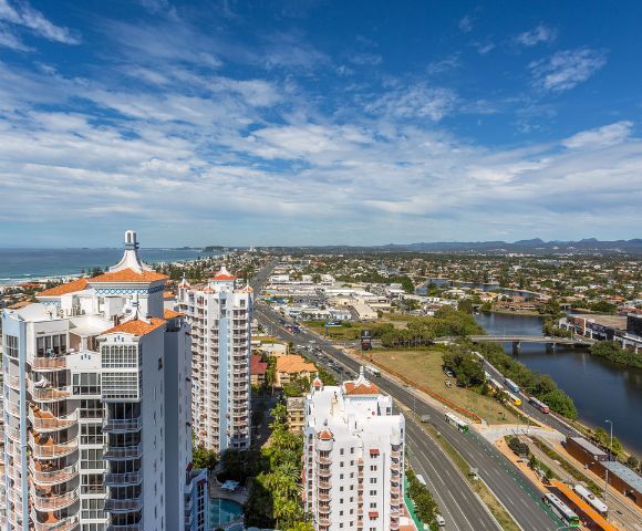 Aerial view of coastal city with modern skyscrapers, a highway, lush greenery, and the ocean under a partly cloudy sky.
