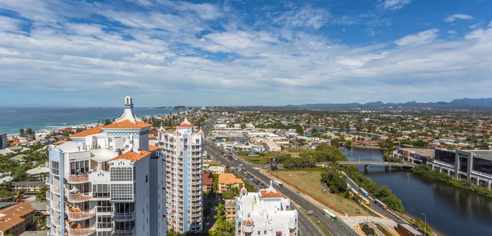 Aerial view of coastal city with modern skyscrapers, a highway, lush greenery, and the ocean under a partly cloudy sky.