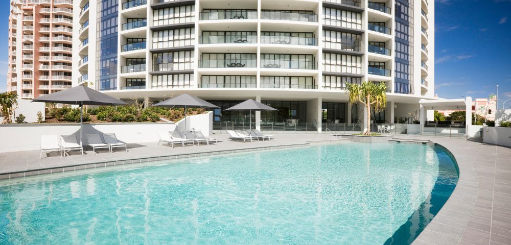 Modern high-rise building behind a large turquoise pool with lounge chairs and umbrellas on a sunny day.