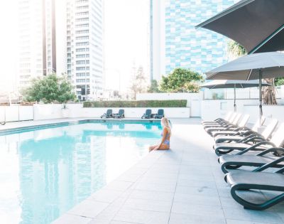 Woman in swimsuit sitting by an outdoor pool with lounge chairs, umbrellas, and tall buildings in the background on a sunny day.