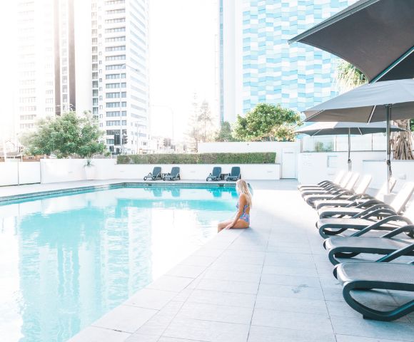 Woman in swimsuit sitting by an outdoor pool with lounge chairs, umbrellas, and tall buildings in the background on a sunny day.