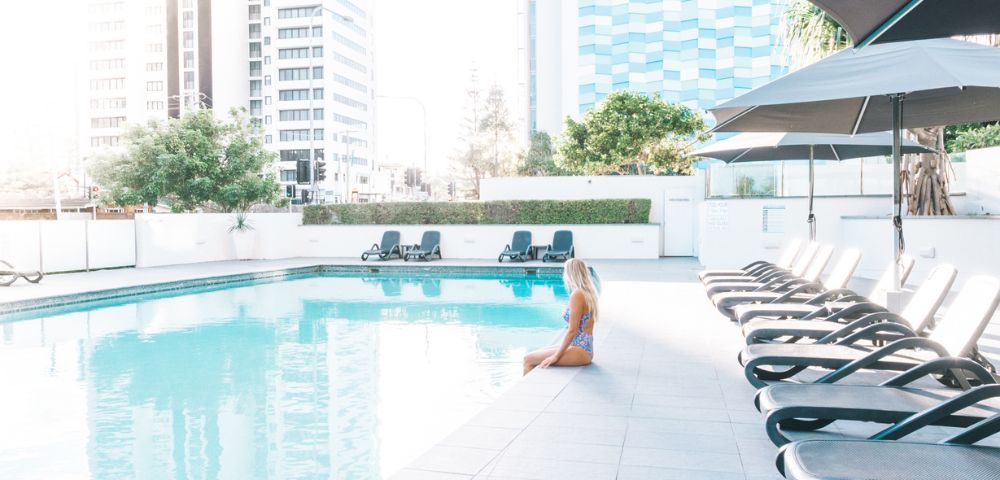 Woman in swimsuit sitting by an outdoor pool with lounge chairs, umbrellas, and tall buildings in the background on a sunny day.