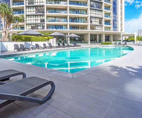 Modern outdoor pool area with black lounge chairs and umbrellas, set in front of a multi-story building with balconies and surrounded by light gray tiles and greenery.