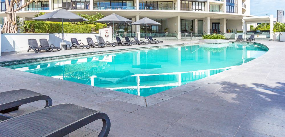 Modern outdoor pool area with black lounge chairs and umbrellas, set in front of a multi-story building with balconies and surrounded by light gray tiles and greenery.
