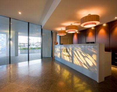 Modern hotel reception area with a white counter, pendant lights, wood-paneled wall, and large glass windows overlooking greenery.