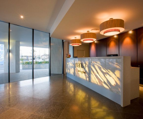 Modern hotel reception area with a white counter, pendant lights, wood-paneled wall, and large glass windows overlooking greenery.