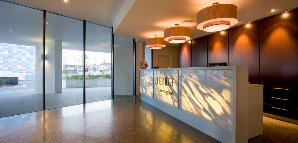 Modern hotel reception area with a white counter, pendant lights, wood-paneled wall, and large glass windows overlooking greenery.