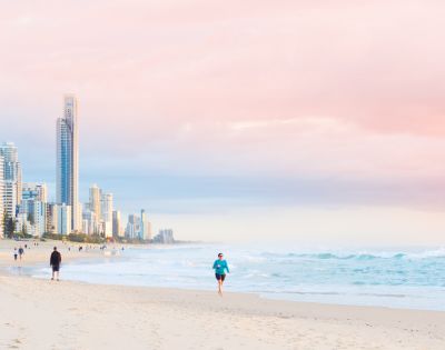 Person jogging along a sandy beach with ocean waves, distant figures, and tall buildings under a pink and blue sunrise or sunset sky.