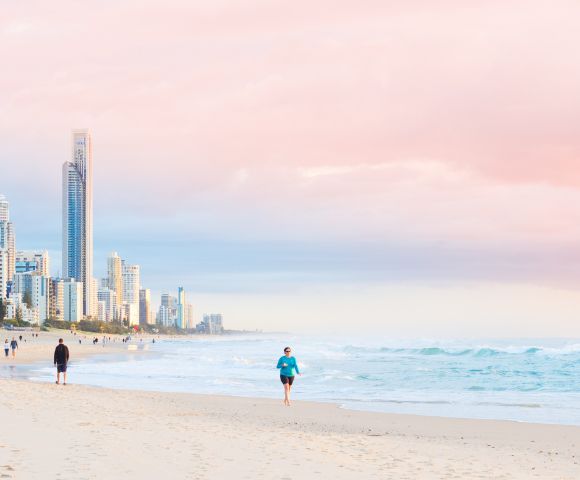 Person jogging along a sandy beach with ocean waves, distant figures, and tall buildings under a pink and blue sunrise or sunset sky.