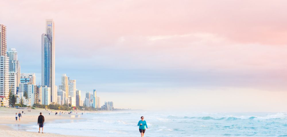 Person jogging along a sandy beach with ocean waves, distant figures, and tall buildings under a pink and blue sunrise or sunset sky.
