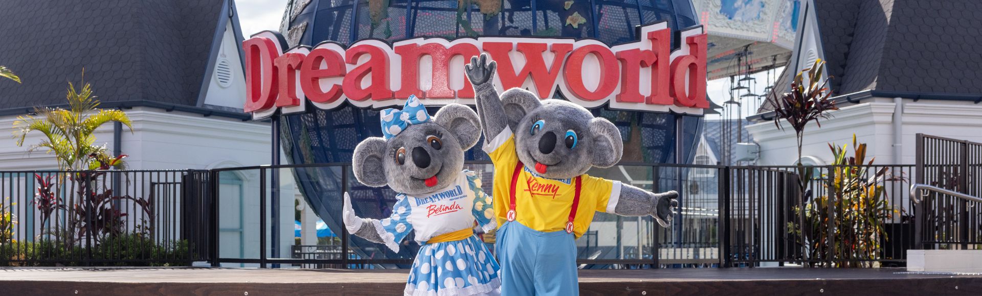 Two koala mascots, dressed in colorful outfits, pose joyfully in front of the Dreamworld sign and globe. Sunny day at the theme park.