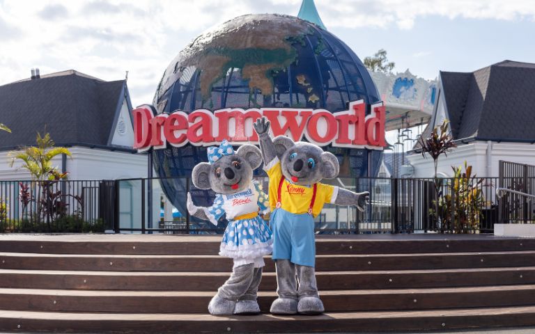 Two koala mascots, dressed in colorful outfits, pose joyfully in front of the Dreamworld sign and globe. Sunny day at the theme park.