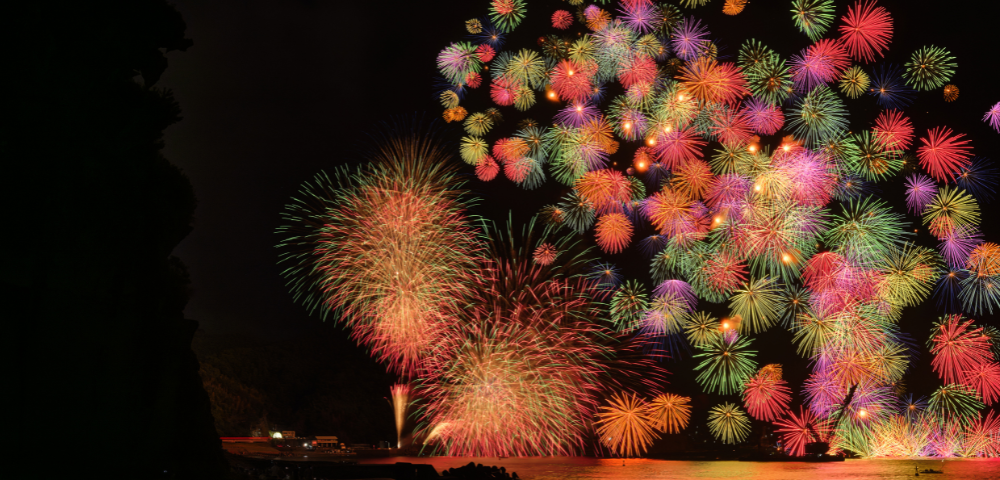 A vibrant display of fireworks lights up the night sky over a beach, with colorful bursts reflecting on the water's surface.