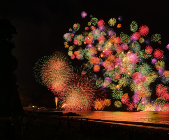 A vibrant display of fireworks lights up the night sky over a beach, with colorful bursts reflecting on the water's surface.